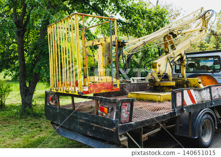 Park maintenance truck with elevated lift, rusted bucket secured on flatbed, folded crane boom, leafy backdrop, soft morning light, equipment staged for arborist repairs Park maintenance truck with elevated lift, rusted bucket secured on flatbed, folded crane boom, leafy backdrop, soft morning light, equipment staged for arborist repairs 140651015