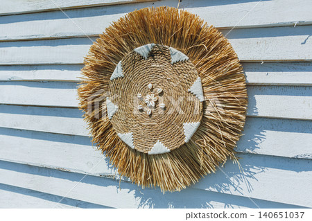 Handmade straw wreath against weathered siding with white accents. Rustic outdoor decoration featuring sunlit straw wreath with geometric details on wooden siding Handmade straw wreath against weathered siding with white accents. Rustic outdoor decoration featuring sunlit straw wreath with geometric details on wooden siding 140651037