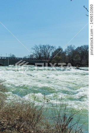 Water flows fiercely at Niagara Falls with a view of the bridge and nearby land during a sunny day 140651060