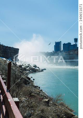 Visitors watch water flow over Niagara Falls in bright sunlight with mist rising from the falls and city buildings in the distance 140651071