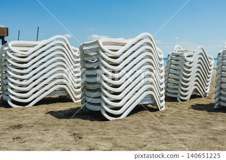 Sunbeds and chairs organized beachside. Arrange sunbathing furniture along sandy coast. Alignment of sunbeds and white chairs prepares for busy summer tourist season Sunbeds and chairs organized beachside. Arrange sunbathing furniture along sandy coast. Alignment of sunbeds and white chairs prepares for busy summer tourist season 140651225