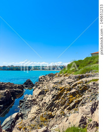 Coastline view showing rocky shore and clear water under bright sky with distant buildings and vegetation in summer season Coastline view showing rocky shore and clear water under bright sky with distant buildings and vegetation in summer season 140651253