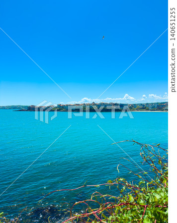 Bright blue sea and clear sky view from the shore with distant buildings along the coastline in a sunny day Bright blue sea and clear sky view from the shore with distant buildings along the coastline in a sunny day 140651255