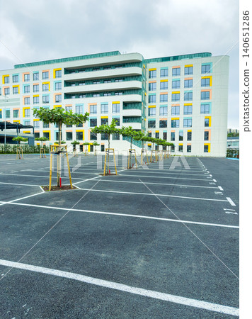 Empty parking lot with young trees in front of a large modern building on a cloudy day in an urban area Empty parking lot with young trees in front of a large modern building on a cloudy day in an urban area 140651286