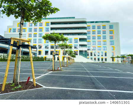 Building stands next to a large parking area with newly planted trees under a cloudy sky in a modern urban setting Building stands next to a large parking area with newly planted trees under a cloudy sky in a modern urban setting 140651301