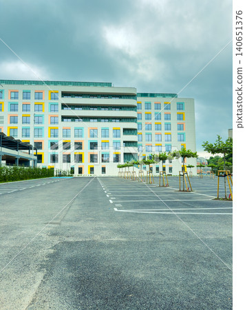 Modern building stands tall in a newly developed area with empty parking spaces and trees under a cloudy sky Modern building stands tall in a newly developed area with empty parking spaces and trees under a cloudy sky 140651376