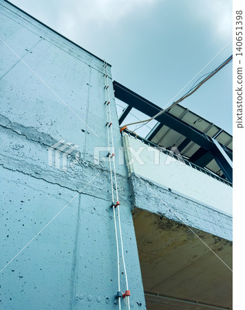 Building construction shows blue concrete wall with white cables leading to upper structure under cloudy sky Building construction shows blue concrete wall with white cables leading to upper structure under cloudy sky 140651398