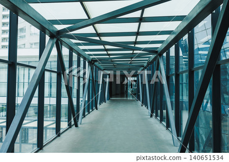 Modern walkway connects buildings in urban area during daytime showing clear glass walls and metal frames Modern walkway connects buildings in urban area during daytime showing clear glass walls and metal frames 140651534