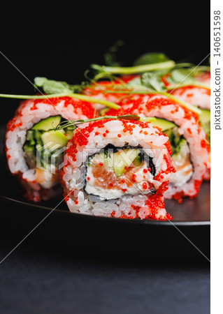 Eating sushi rolls with cucumber and fish on a black plate in a dimly lit setting with green toppings on top Eating sushi rolls with cucumber and fish on a black plate in a dimly lit setting with green toppings on top 140651598