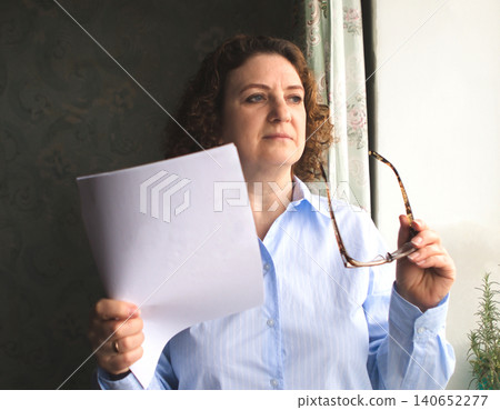 Mature woman holding document and glasses looking thoughtful by a window in soft natural light Mature woman holding document and glasses looking thoughtful by a window in soft natural light 140652277