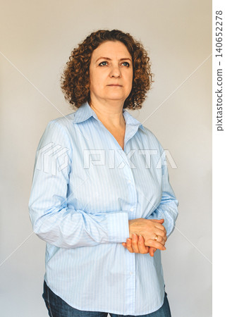 Thoughtful middle-aged woman in blue shirt standing with folded hands for portrait Thoughtful middle-aged woman in blue shirt standing with folded hands for portrait 140652278