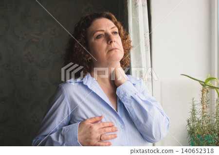 Middle-aged woman looking concerned while holding her chest and neck by a window 140652318