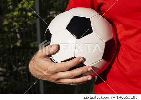 Man in a red T shirt holding a black and white soccer ball outdoors, reflecting a connection to the global soccer World cap. 140653185
