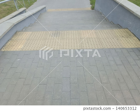 Top down aerial view of a paved sloping walkway with yellow tactile paving strips, handrails on both sides, and green grass bordering the path... 140653312