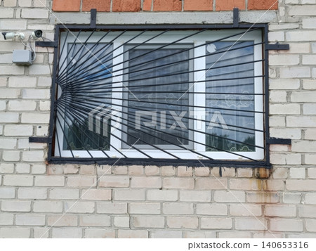A white two-lens security camera mounted on a brick wall above a window with heavy black metal bars and grate. The building exterior shows signs of aging, creating a surveillance-focused urban scene A white two-lens security camera mounted on a brick wall above a window with heavy black metal bars and grate. The building exterior shows signs of aging, creating a surveillance-focused urban scene 140653316