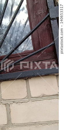 A weathered window with a patterned white curtain and diagonal black security bars, peeling wooden frame below, and a brick wall base. An urban detail shot focused on protection and decay... 140653317