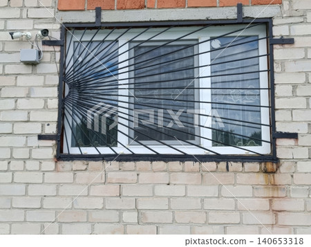 A white two-lens security camera mounted on a brick wall above a window with heavy black metal bars and grate. The building exterior shows signs of aging, creating a surveillance-focused urban scene A white two-lens security camera mounted on a brick wall above a window with heavy black metal bars and grate. The building exterior shows signs of aging, creating a surveillance-focused urban scene 140653318