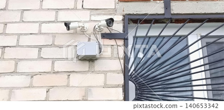Two white security cameras mounted on a white brick wall next to a window with a decorative metal grate, connected to a grey electrical box.. 140653342