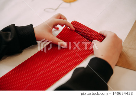 Child rolling red beeswax candle sheet by hand, cozy DIY craft process indoors Child rolling red beeswax candle sheet by hand, cozy DIY craft process indoors 140654438
