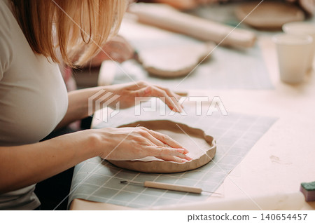 close-up female hands make a clay plate, align the shape with special tools in the workshop close-up female hands make a clay plate, align the shape with special tools in the workshop 140654457