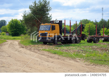 Yellow timber truck with crane arm parked near dirt road, empty log carrier semi trailer on green glade landscape 140654705