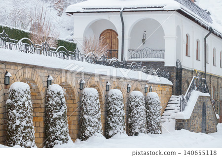 A picturesque scene of a European architectural campus courtyard blanketed in soft snow, featuring stone walls and manicured topiaries 140655118