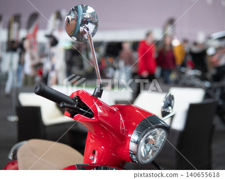 Red Scooter Front In Showroom, Glossy Chrome Mirror And Headlight In Sharp Focus, Blurred Shoppers And Display Booths In Background, Modern Lifestyle Composition With Retail Lighting 140655618