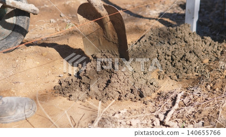 Shovel Pouring Wet Concrete Onto Plywood Near Wheelbarrow And Boots, Dusty Sunlight Highlights Textured Slurry And Scattered Soil, Manual Mixing For Small Backyard Repair 140655766