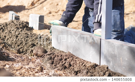 Mason Aligning Concrete Curb With Stringline And Measurement, Blocks Being Leveled Into Trench On Prepared Base, Gravel And Soil Visible, Hands With Gloves Guiding Layout For Urban Edging Mason Aligning Concrete Curb With Stringline And Measurement, Blocks Being Leveled Into Trench On Prepared Base, Gravel And Soil Visible, Hands With Gloves Guiding Layout For Urban Edging 140655808