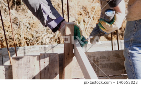 Hands Assembling Wooden Construction Formwork For Foundation, Workers Aligning Timber And Securing Rebar With Gloves And Drill At Muddy Excavation, Temporary Braces And Concrete Forms 140655828