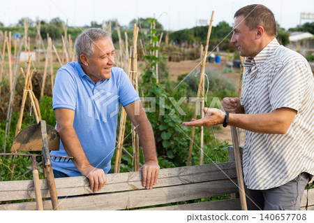 Two male farmers talking outside next to wooden fence on background with farm Two male farmers talking outside next to wooden fence on background with farm 140657708