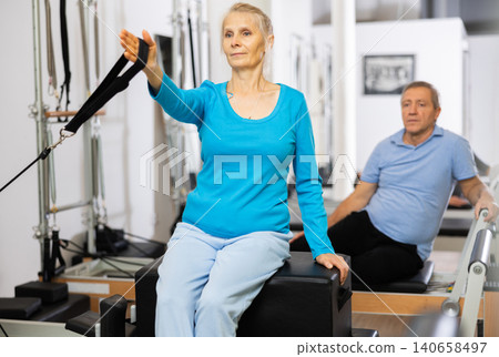 Mature couple using bed of Pilates reformer bed equipment while training together in wellness center. Wellbeing and healthy lifestyle concept Mature couple using bed of Pilates reformer bed equipment while training together in wellness center. Wellbeing and healthy lifestyle concept 140658497