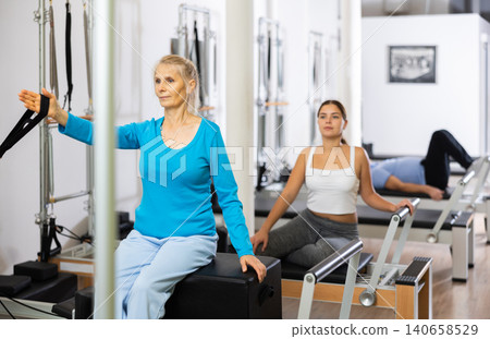 Mature woman in sportswear sitting and doing Pilates exercises for hands on reformer bed equipment in fitness center Mature woman in sportswear sitting and doing Pilates exercises for hands on reformer bed equipment in fitness center 140658529