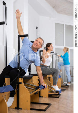 Focused mature man performing stretching exercises while using pilates Wanda chair machine in rehabilitation center Focused mature man performing stretching exercises while using pilates Wanda chair machine in rehabilitation center 140658690