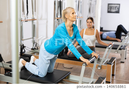 Positive aged woman doing Pilates exercises for lower back while lying on reformer bed in rehabilitation center. Fitness and wellness concept Positive aged woman doing Pilates exercises for lower back while lying on reformer bed in rehabilitation center. Fitness and wellness concept 140658861