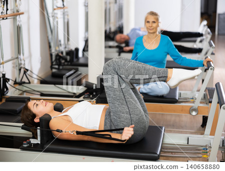 Young woman lying down on bed of Pilates reformer machine bed, with legs up, working out and training at gym studio Young woman lying down on bed of Pilates reformer machine bed, with legs up, working out and training at gym studio 140658880
