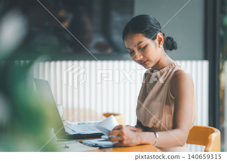 Concerned Asian businesswoman reviewing financial papers beside laptop in modern cafe office, showing budget problems, accounting pressure, data analysis, business risk, and difficult decisions. Concerned Asian businesswoman reviewing financial papers beside laptop in modern cafe office, showing budget problems, accounting pressure, data analysis, business risk, and difficult decisions. 140659315