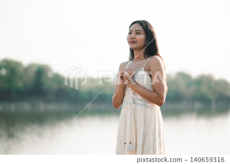 Young Asian woman in a white dress standing by a calm lake with hands clasped, soft daylight and copy space, showing gratitude, hope, peaceful nature, outdoor wellness, and mindful travel. Young Asian woman in a white dress standing by a calm lake with hands clasped, soft daylight and copy space, showing gratitude, hope, peaceful nature, outdoor wellness, and mindful travel. 140659316