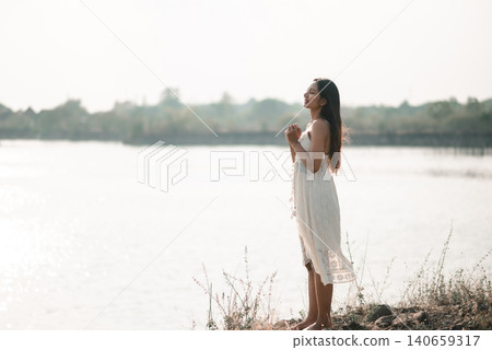 Young Asian woman in a white dress standing on a lakeshore with hands clasped in soft daylight, showing prayer, gratitude, hope, peaceful nature, mindful breathing, wellness, and spiritual calm. Young Asian woman in a white dress standing on a lakeshore with hands clasped in soft daylight, showing prayer, gratitude, hope, peaceful nature, mindful breathing, wellness, and spiritual calm. 140659317