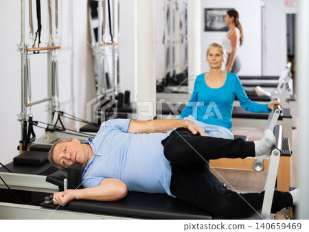 Focused mature male pensioner doing Pilates exercise for back on reformer bed equipment in rehabilitation center Focused mature male pensioner doing Pilates exercise for back on reformer bed equipment in rehabilitation center 140659469