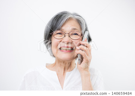 A bespectacled senior woman smiles and communicates on her smartphone against a white background. Hello? 140659484