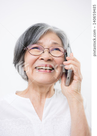 A bespectacled senior woman smiles and communicates on her smartphone against a white background. Hello? A bespectacled senior woman smiles and communicates on her smartphone against a white background. Hello? 140659485
