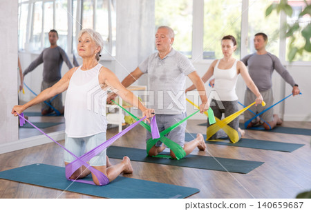Elderly woman performs exercises with elastic band during Pilates strength training, kneeling Elderly woman performs exercises with elastic band during Pilates strength training, kneeling 140659687