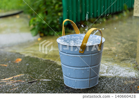 Rainwater Harvesting System Collecting Roof Runoff into Bucket for Sustainable Water Conservation. Rainwater flows from a roof gutter and drainage system into a metal bucket for collection and future Rainwater Harvesting System Collecting Roof Runoff into Bucket for Sustainable Water Conservation. Rainwater flows from a roof gutter and drainage system into a metal bucket for collection and future 140660428