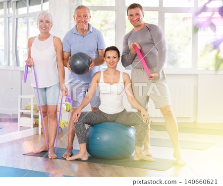 Group of women and men of different ages in sportswear with fitness balls and resistance bands posing together in sports studio, ready for training Group of women and men of different ages in sportswear with fitness balls and resistance bands posing together in sports studio, ready for training 140660621