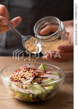 Woman hand pouring roasted sesame seed into a bowl with cucumber to making Korean cucumber salad Woman hand pouring roasted sesame seed into a bowl with cucumber to making Korean cucumber salad 140661027