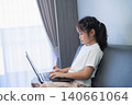 Young girl sitting on sofa using a laptop computer near window with curtains, focused on screen, casual attire, home interior background, natural lighting 140661064