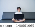 Young girl sitting on a modern gray sofa cross-legged, using a black laptop with a plain white wall background, demonstrating focus and digital technology use 140661065