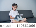 Young girl with long dark hair sitting on a modern sofa, engaging with a tablet device in a minimalistic living room, emphasizing technology use among children 140661066