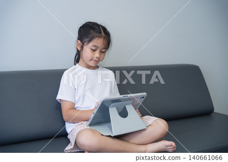 Young girl with long dark hair sitting on a modern sofa, engaging with a tablet device in a minimalistic living room, emphasizing technology use among children Young girl with long dark hair sitting on a modern sofa, engaging with a tablet device in a minimalistic living room, emphasizing technology use among children 140661066